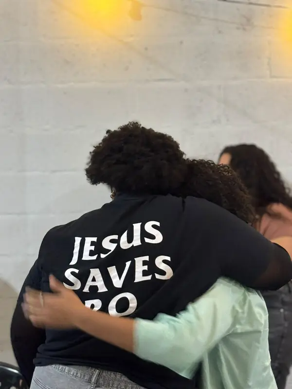 Duas pessoas se abraçam num encontro da igreja; nas costas de uma delas, a camiseta diz “Jesus saves”.