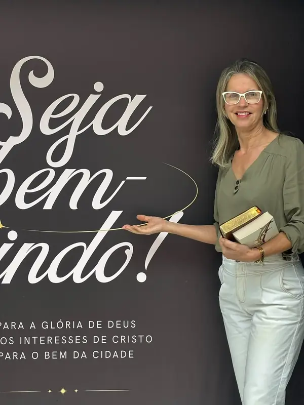 Uma mulher sorri e aponta para o painel “Seja bem-vindo!” da igreja, com uma Bíblia na mão.
