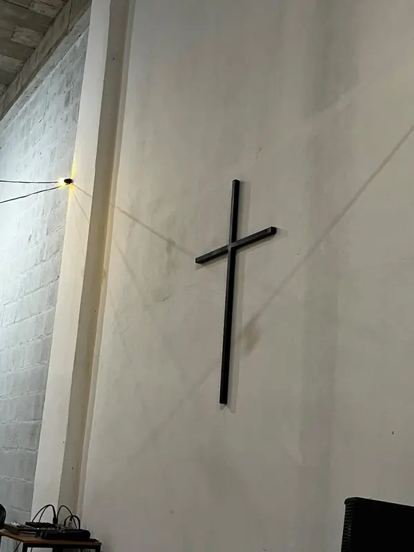 A cruz na parede branca do salão, iluminada pela luz quente de uma lâmpada presa ao pilar.