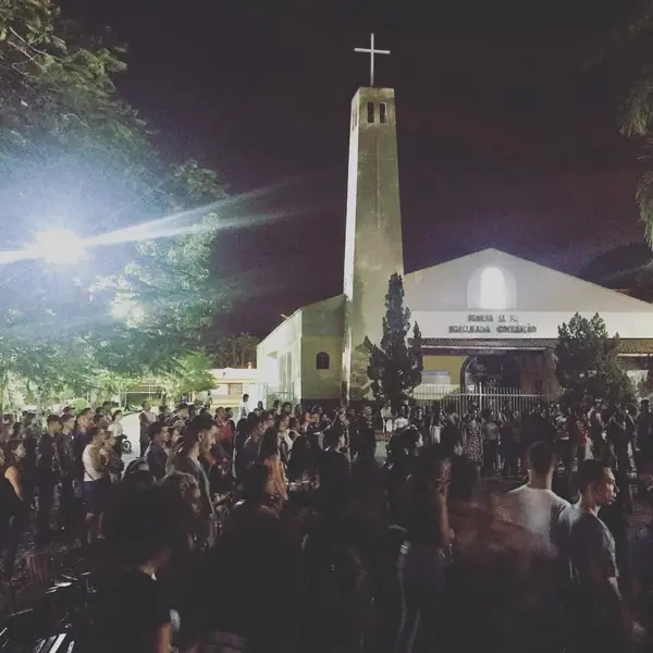 Uma multidão reunida à noite numa praça, diante de uma igreja de torre alta com uma cruz no alto.